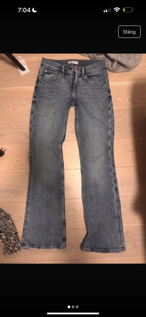 Blå bootcut jeans med fickdetalj - Säljer ett par blå bootcut jeans med klassisk femficksmodell och extra ficka på benet. Jeansen har en snygg tvättad look och raka ben som slutar i en lätt utsvängning. Perfekta för en avslappnad och trendig stil.