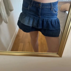 Mörkblå jeansskort med volang - Säljer en mörkblå jeansskort med volangkant och klassiska fickor både fram och bak. Skorten har en feminin och trendig look med hög midja och knappgylf. Perfekt för dig som gillar denim och vill ha något bekvämt men ändå snyggt.