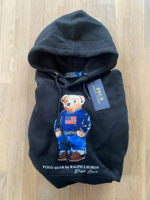 Svart hoodie Polo Bear Ralph Lauren - Svart hoodie från Ralph Lauren med ikoniska Polo Bear-trycket framtill. Hoodien har huva med dragsko, magficka och ribbade muddar. Materialet är mjukt och skönt, perfekt för en chill och stilren look. Märkeslapp kvar på plagget.