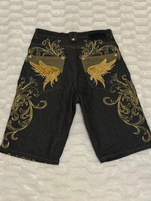 Jorts med brodyr - Unika svarta baggy ”jorts” jeansshorts med detaljerad guld brodyr och stora vingmotiv på bakfickorna🪽, köpte dom på secondhand och har ingen aning om vilket märke det är, men dom är i gott skick och riktigt snygga ✨dom är i storlek 34/36, jag har 34 men dom här var lite större men passade ändå bra i midjan. För fler bilder är det bara att skriva till mig så löser jag det!