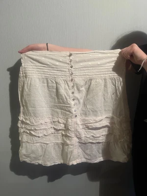 Vit kort klänning med volanger - Söt vit kort klänning med knappar framtill och volangdetaljer. Klänningen har ett diskret prickigt mönster och är tillverkad i ett lätt, luftigt tyg som passar perfekt till varmare dagar. Smickrande passform med fina detaljer upptill. Finns liten slitning vid längst upp där armarna är 