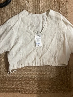 Vit croppad linnetopp med v-ringning - Snygg vit croppad topp i linne med v-ringning och korta ärmar. Toppen har dragsko i nederkanten för justerbar passform och en avslappnad, luftig stil. Perfekt för varma dagar och enkel att matcha med jeans eller kjol.