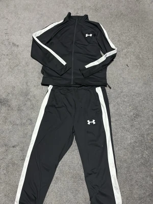 Svart träningsset från Under Armour - Säljer ett svart träningsset från Under Armour med vita ränder längs ärmar och byxben. Setet består av ziptröja och byxor i mjukt, stretchigt material. Perfekt för gymmet eller löprundan. Klassisk logga på byxan och dragkedja på tröjan.