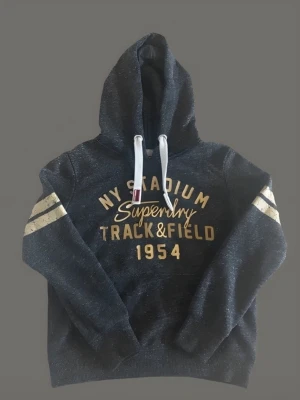Mörkblå Superdry hoodie med tryck - Snygg mörkblå hoodie från Superdry med gul text 'NY Stadium Track & Field 1954' på bröstet och vita ränder på ärmarna. Huvan har vita dragsnören och insidan är mjuk. Perfekt för en sportig och avslappnad stil.