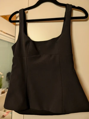 Svart linnetopp med rosett - Stilren svart linnetopp med breda axelband och en snygg rosettdetalj i ryggen. Toppen har en lätt utsvängd passform och är perfekt för att skapa en chic look. Materialet känns mjukt och följsamt mot kroppen. 