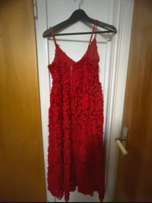 Röd långklänning med 3D-blommor - Snygg röd långklänning med tunna axelband och v-ringning. Klänningen har ett unikt 3D-blommigt mönster över hela tyget som ger en lyxig och trendig känsla. Perfekt för dig som vill sticka ut och älskar detaljer. Materialet känns lätt och luftigt.
