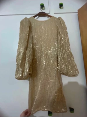 Glittrande guldig paljettklänning - Supersnygg guldig klänning täckt av paljetter med puffiga långärmar och rak passform. Klänningen har rund halsringning och är perfekt för dig som vill sticka ut. Materialet är glittrigt och ger en lyxig känsla.