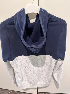 Blå och grå hoodie med dragkedja - Snygg hoodie från Denim Goods i tvåfärgad design med mörkblå ovandel och ljusgråmelerad nederdel. Dragkedja framtill, fickor och tryck på bröstet med texten 'ATHL DEPT'. Mjuk bomullsmix och huva med dragsko för extra stilpoäng. Varje kläder koster 250 st 