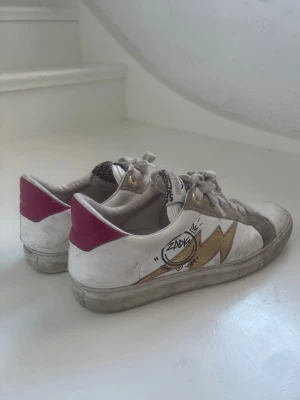 Zadig skor - Unika vita sneakers med en stor guld-blixt och grafiskt tryck på sidan. Skorna har röda detaljer vid hälen och vita snören. Tillverkade i läder med klassisk låg siluett och platt sula. Perfekta för dig som vill sticka ut med coola detaljer.