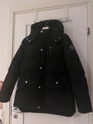 Svart Moose Knuckles 3Q jacka  - Snygg svart dunjacka från Moose Knuckles med huva, silverfärgade knappar och dragkedja. Jackan har två stora fickor framtill och logga på ärmen. Perfekt för kalla vinterdagar och riktigt varm tack vare dunfyllningen. Modellen kommer utan päls. Köptes för ungefär 3500kr för 4 år sen och jackan är limited edition! XL i barnstorlek=Xxs-Xs-herr kolla 5te bild! Jackan är äkta