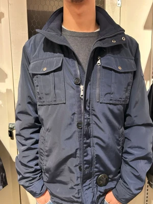  Mörkblå J Lindeberg Fieldjacket - Snygg mörkblå Bailey Poly Jacket vindjacka med hög krage och dragkedja samt knappar framtill. Jackan har två stora bröstfickor med lock och tryckknappar. STORLEK L, NYPRIS 3400kr!