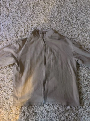 Beige hoodie med dragkedja - En stilren beige hoodie med hel dragkedja framtill och hög krage. Hoodien har en minimalistisk design utan synliga loggor eller tryck och är tillverkad i mjukt bomullsliknande material. Perfekt för en clean och avslappnad look.