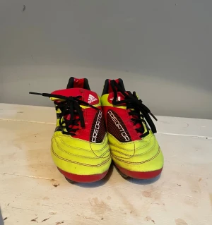 Adidas Predator gula fotbollsskor - Säljer ett par klassiska Adidas Predator fotbollsskor i gul och röd färg med svarta detaljer och snörning. Skorna har de ikoniska tre svarta ränderna på sidan och Predator-loggan på plösen. Yttermaterialet är syntet och sulan har dobbar för bra grepp på planen.