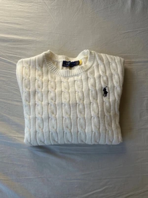 Vit kabelstickad tröja Polo Ralph Lauren - Kabelstickad vit tröja från Polo Ralph Lauren med rund halsring och klassisk broderad logga på bröstet. Tröjan har en mjuk och skön känsla, perfekt för dig som gillar tidlösa och stilrena outfits.