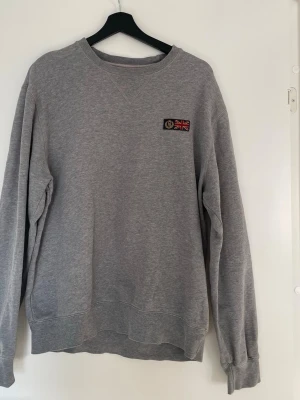Henri Lloyd sweatshirt – grå – M - Grå sweatshirt från Henri Lloyd med klassisk design och broderad patch på bröstet.  ✔ Storlek: M ✔ Färg: Grå ✔ Modell: Sweatshirt ✔ Skick: Använd – mindre slitage och små hål finns (se bilder