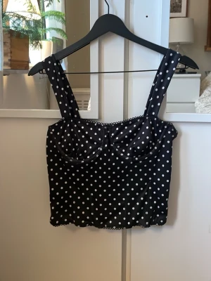 Svart prickig bustier-top - Snygg svart bustier-top med vita prickar och breda axelband. Toppen har markerade kupor och är gjord i ett glansigt, mjukt material som ger en retrovibe. Perfekt att styla med jeans eller kjol för en trendig look. I storlek 36 