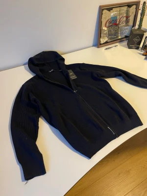 Mörkblå stickad zip hoodie - Säljer en mörkblå hoodie från Oscar Jacobson med dragkedja framtill och ribbad struktur. Hoodien har en klassisk huva och är tillverkad i ett mjukt, stickat material som känns riktigt skönt. Perfekt för dig som gillar stilrena och bekväma plagg. Helt ny i storlek medium.