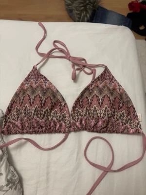 Mönstrad rosa bikiniöverdel med knyt - Säljer en snygg trekantsbikini i rosa och beige toner med ett unikt mönster. Bikinin har knyt i nacken och ryggen för justerbar passform. Perfekt för sommaren och strandhäng! Storlek L, men passar folk med D/E kupa. Använd en gång endast, annars har jag bara testat den. Aldrig badat med den.