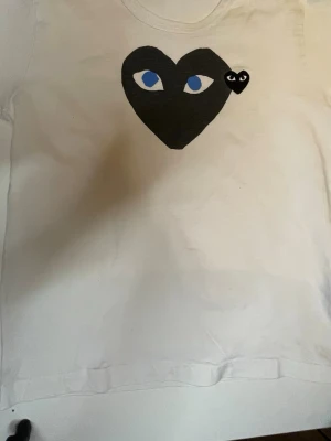 Vit Comme des Garçons (CDG) T-shirt  - Säljer en vit t-shirt från Comme des Garçons PLAY med det ikoniska svarta hjärtat med blå ögon tryckt på bröstet och en liten svart hjärtdetalj bredvid. Klassisk rund hals och korta ärmar. Pris kan diskuteras vid snabbaffär. Tveka på att ställa frågor 