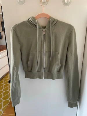 Grön croppad hoodie med dragkedja - Snygg croppad hoodie i ljusgrönt med dragkedja framtill och två fickor. Tröjan har huva med dragsnören och ribbade muddar vid ärmslut och nederkant. Perfekt för en avslappnad och trendig look.