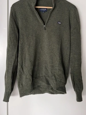 Olivgrön halvzip från Lexington - Olivgrön långärmad tröja från Lexington med halv dragkedja och liten logotyp på bröstet. Tröjan har ribbade muddar vid ärmslut och nederkant samt en klassisk, avslappnad passform. Perfekt för lager på lager och snygg till jeans.