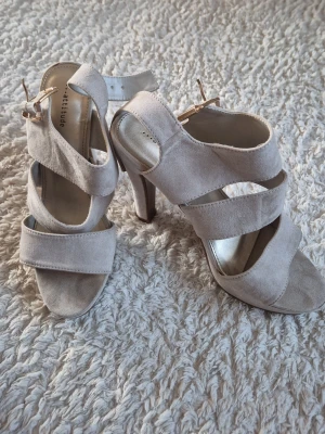 Beige mocka-pumps med klack - Säljer ett par snygga pumps från Attitude i beige mocka. Skorna har öppen tå, breda remmar och hög klack. Stängs med spänne vid ankeln. Perfekta för dig som vill ha en stilren och trendig look.