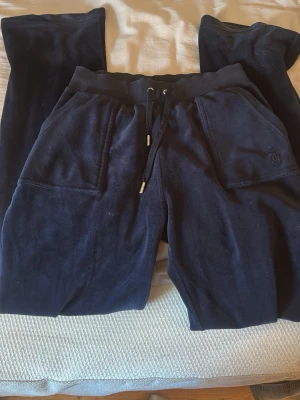 Mörkblå velour mjukisbyxor Juicy Couture - Mörkblå mjukisbyxor i mjuk velour från Juicy Couture. Byxorna har bred resår i midjan med dragsko, stora fickor fram och bakfickor med knapp. Perfekta för chill dagar och har en klassisk relaxed fit. Dessa byxor är i bra längd allt silver är kvar