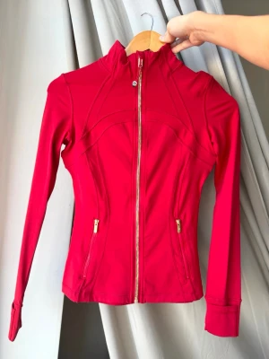 Röd träningsjacka - Snygg röd träningsjacka från Lululemon med figurnära passform, hel dragkedja framtill och två fickor med guldfärgade dragkedjor. Tillverkad i stretchigt funktionsmaterial som andas. Perfekt för gymmet eller löprundan.