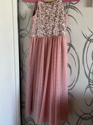 Rosa långklänning med paljetter - Elegant rosa långklänning med glittriga paljetter för barn i 7 års åldern.  Klänningen är ärmlös och har en mjuk, romantisk look som passar perfekt till speciella tillfällen.