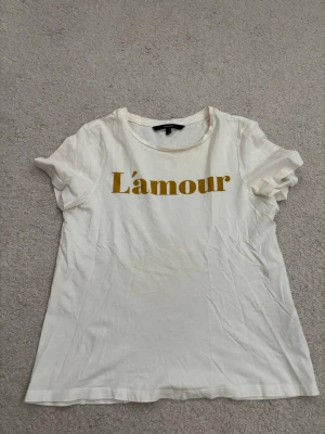 Vit t-shirt med L'amour-tryck - Snygg vit t-shirt med gul text 'L'amour' på bröstet. Klassisk rund halsringning och korta ärmar. Perfekt till jeans eller kjol för en avslappnad look. Tillverkad i mjuk bomull som känns skön mot huden.