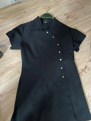 Svart kort klänning med knappar - Snygg svart kort klänning med asymmetrisk knäppning framtill och ståkrage. Klänningen har korta ärmar och en figurnära passform. Lämpar flr sfgörs kläder eller frisör eller hudterapeut Perfekt för dig som gillar minimalistisk och stilren design.