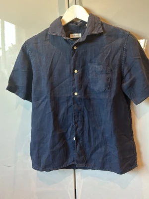 Mörkblå linneskjorta från Gant - Kortärmad skjorta från Gant i mörkblått linne. Klassisk krage, bröstficka och vita knappar framtill. Perfekt för en avslappnad och stilren look under varmare dagar.