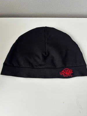 Syna skull cap - Snygg svart mössa med en cool röd broderad logga framtill. Tillverkad i mjukt och stretchigt material som sitter skönt på huvudet. Perfekt accessoar för att hålla stilen när det är kallt ute.