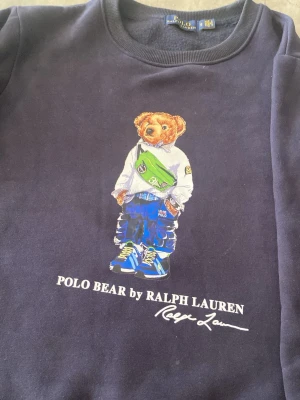 Mörkblå Polo Bear sweatshirt Ralph Lauren - Säljer en mörkblå sweatshirt från Ralph Lauren med den ikoniska Polo Bear-trycket framtill. Tröjan har rund halsringning och ribbade muddar. Tillverkad i mjukt material som känns skönt mot huden. Perfekt för dig som gillar streetwear och klassisk stil.