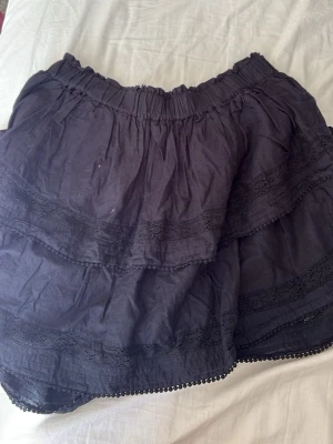 Svart volangkjol med spetsdetaljer - Kort svart kjol med volanger och fina spetsdetaljer. Kjolen har resår i midjan för skön passform och är perfekt för dig som gillar en söt och enkel stil. Materialet känns lätt och passar bra till många olika outfits.