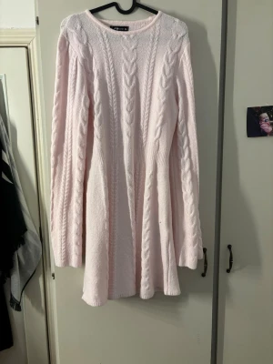 Ljusrosa stickad klänning - Supersöt ljusrosa stickad klänning med kabelstickat mönster längs ärmar och framtill. Klänningen har rund halsringning, lång ärm och lätt utställd kjol. Perfekt för dig som gillar mjuka och feminina plagg.