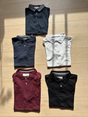 5-pack Pikétröjor från Pier One - Fem pikétröjor från Pier One. En svart, en mörkblå, en vit, en vinröd och en mörkblå med mönster och grå krage. Alla tröjor har krage och knappar framtill.  70kr styck eller alla för 300kr