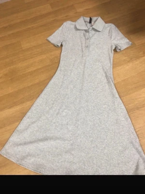 Ljusgrå kortärmad pikéklänning - En enkel och stilren ljusgrå klänning med pikékrage och knappar framtill. Klänningen har korta ärmar och är tillverkad i ett mjukt bomullsmaterial. Passformen är normal och längden går lite över knäna. Perfekt för en avslappnad och bekväm look. Finns små smink fläckar (se bild) använd 1 gång