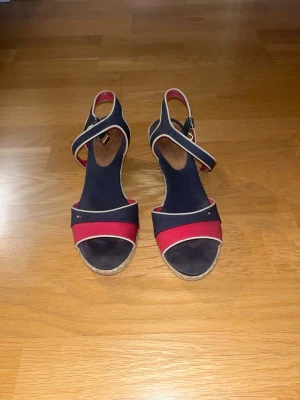 Tommy Hilfiger sandaler kilklack storlek 39 - Hilfiger i klassiska färger marinblått, rött och vitt. De är i bra skick med normalt slitage, men fortfarande väldigt fina och fullt användbara. Sandalerna har en bekväm kilklack som gör dem sköna att gå i, samt en justerbar rem med spänne för bra passform. Perfekta till sommaren och passar lika bra till vardag som till lite finare tillfällen.