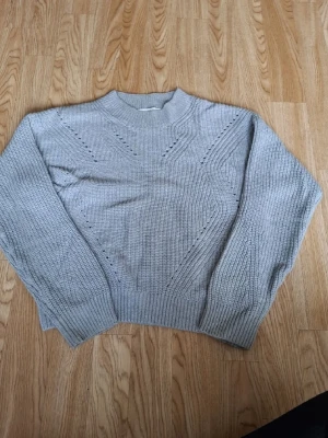 Ljusgrå stickad tröja med mönster - Säljer en ljusgrå stickad tröja med diskret hålmönster framtill och ribbade muddar vid ärmslut och nederkant. Tröjan har en rak passform, hög hals och är perfekt för kyliga dagar. Materialet känns mjukt och bekvämt.