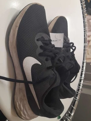 Svarta Nike sneakers med vit sula - Svarta sneakers från Nike med vit sula och stor vit swoosh-logga på sidan. Ovandelen är i meshmaterial för bra andningsförmåga och skon har klassisk snörning. Perfekta för en sportig och avslappnad stil.