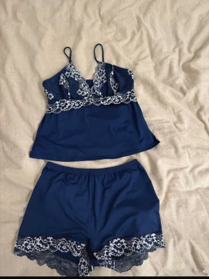 Mörkblå spetsdetaljerat pyjamasset - Sött tvådelat pyjamasset i mörkblått med vita spetsdetaljer. Setet består av ett linne med tunna axelband och spets vid bysten samt ett par shorts med elastisk midja och matchande spets längst ner. Perfekt för en mysig kväll hemma.