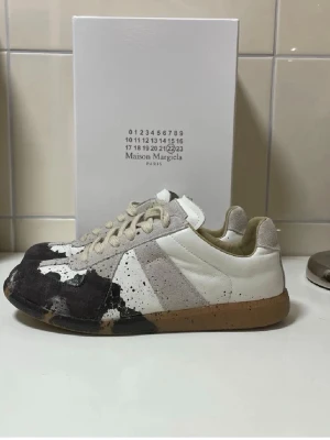 Maison Margiela  - Maison Margiela i vitt och beige läder och mocka med unikt mörkblått färgstänksmönster. Klassisk låg modell med beige snörning och gummisula. Perfekta för dig som gillar statement-plagg och streetwear-vibe.