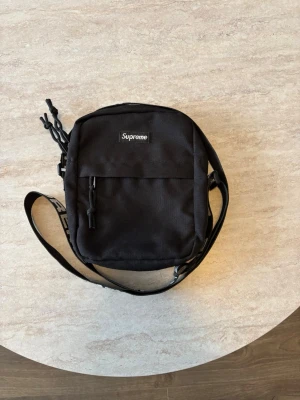 Svart axelväska från Supreme - Supreme axelväska i svart slitstark canvas med justerbar axelrem där Supreme-loggan är tryckt i vitt. Väskan har en fyrkantig form, dragkedjeförsedda fack och minimalistisk design med Supreme-brodyr på framsidan. Perfekt för att bära det viktigaste.