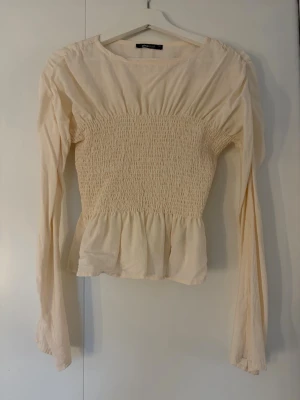 Ljusbeige smockad blus med volang - Supersöt ljusbeige blus med smockad midja och volang nedtill. Långa ärmar och rund halsringning. Liten defekt på höger sida men inget som syns mycket. 