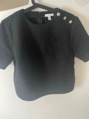 Svart topp med dekorativa knappar H&M - Stilren svart topp från H&M i strukturerat tyg med korta ärmar och rund halsringning. Tre silvriga dekorativa knappar på axeln. Storlek , nyskick