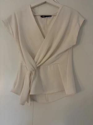 Krämvit omlottopp från Zara - Snygg krämvit omlottopp från Zara med knytning i midjan och korta ärmar. Öppnar med dragkedja på sidan. Den har en liten defekt över högra bröstet men inget som syns mycket. 