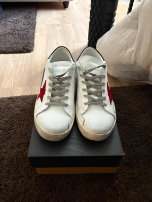 Vita sneakers med röd stjärna - Snygga vita sneakers med en röd stjärna på sidan och grå skosnören. Skorna har svart detalj på hälen och är tillverkade i läder. Perfekta för dig som vill ha en clean look med en cool detalj.