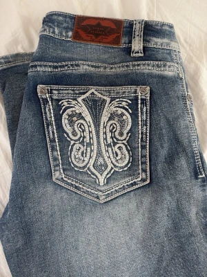 Blåa bootcut jeans med detalj - Säljer ett par blåa bootcut jeans med snyggt broderad bakficka i vitt och silver. köpta för 1200kr