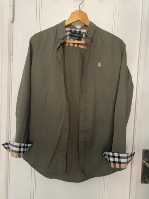 Burberry London skjorta khaki Nova Check-detalj herr L - Snygg Burberry London-skjorta i storlek L — olivgrön/khaki med ikoniskt Nova Check-foder i krage samt ärm och broderad logga på bröstet.  Detaljer: 	•	Modell: Oxford/casual skjorta 	•	Storlek: L (personen i bild är ca 185 cm, 74 kg) 	•	Färg: Khaki/olivgrön                                                          •	Passform: Regular fit 	•	Material: Bomull Skick: Gott begagnat skick — inga synliga fläckar eller skador. Skrynklorna beror på nytvätt. Nypris: ca 2500–3500 kr  